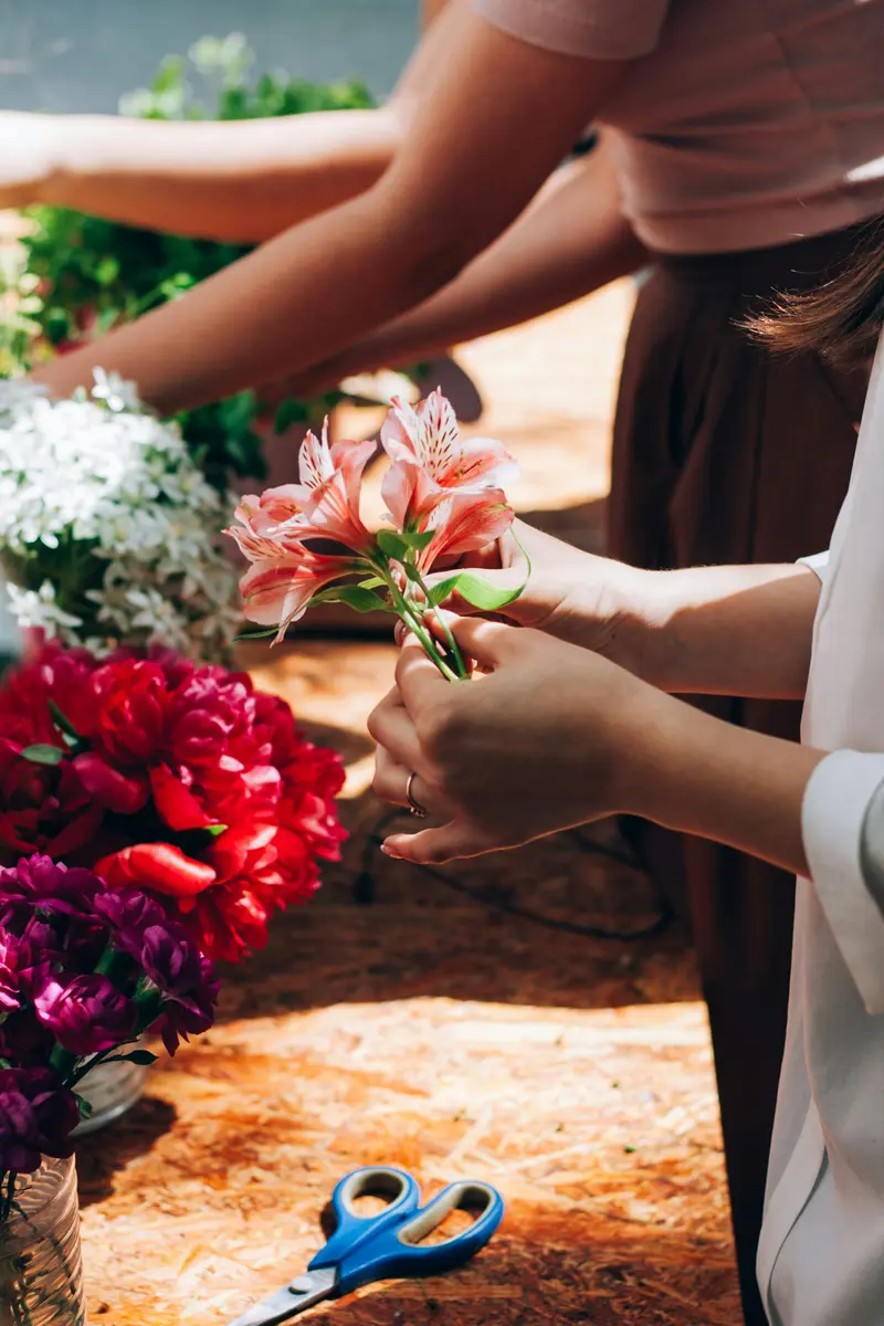 机いっぱいに赤や白の花材が広がり、床に青い持ち手のはさみが置かれている。数人の手が花を選んでいる